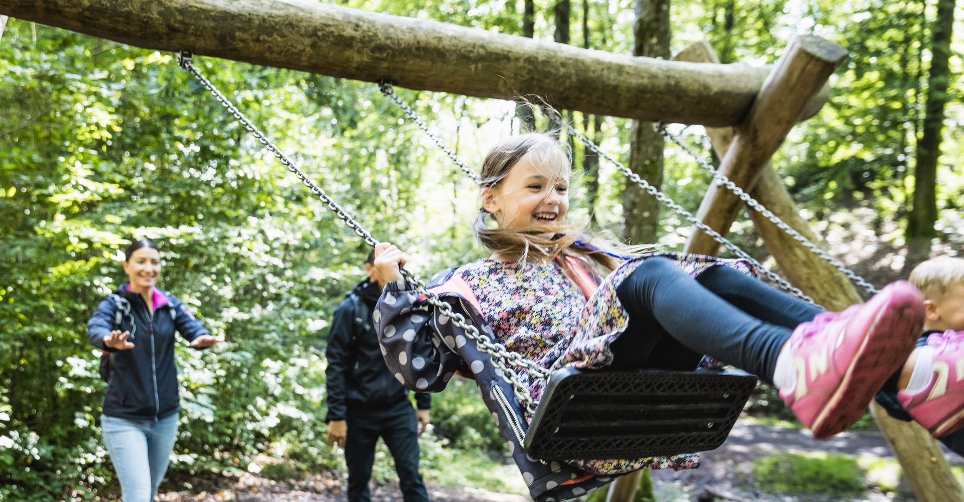 Im Vordergrund ist ein Mädchen mit langen Haaren zu sehen. Sie trägt Zöpfe und lacht, da sie schaukelt. Im Hintergrund steht eine Frau, die sie anschubst. Die Schaukel befindet sich im grünen Laubwald.