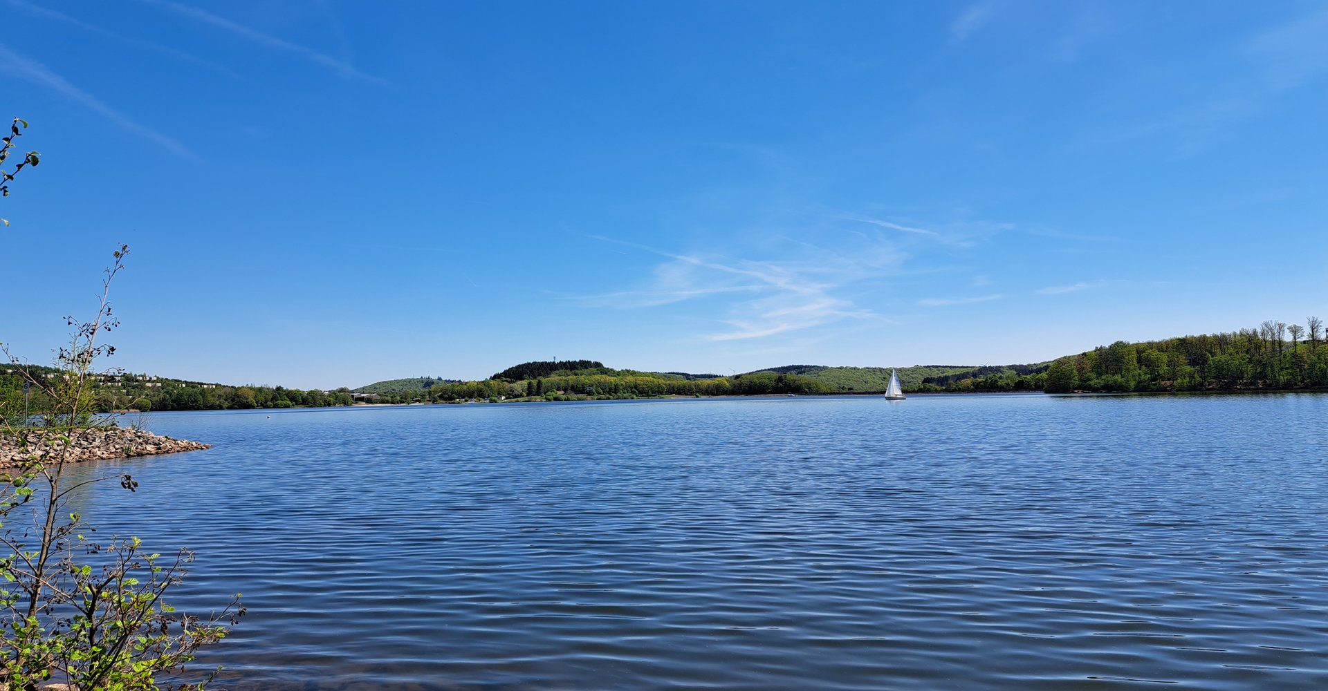 Es ist ein See zu sehen mit grün bewaldetem Ufer. Auf dem See ist ein Segelschiff.