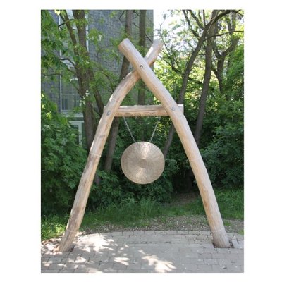 Es sind drei Holzpfosten zu sehen, die a-förmig aufgestellt sind. Daran hängt eine messingfarbene Klangschale.